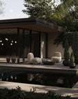 Modern outdoor pavilion with seating area by a pond, surrounded by trees.