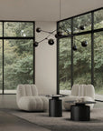 Modern living room with gray armchairs, black tables, and a decorative chandelier in front of large windows with a view of trees.