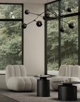 Modern living room with gray armchairs, black tables, and a decorative chandelier in front of large windows with a view of trees.