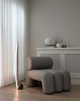 Modern gray armchair in a minimalistic room with light-colored curtains and wooden floor.