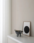 Modern interior with a dark grey colour bowl and chopsticks on a white surface, next to a framed artwork with a black circle on a beige wall from 101 Copenhagen at Spacio retail stores in Mumbai & Kolkata.