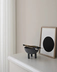 Modern interior with a dark grey colour bowl and chopsticks on a white surface, next to a framed artwork with a black circle on a beige wall from 101 Copenhagen at Spacio retail stores in Mumbai & Kolkata.