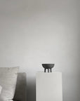 Dark grey colour bowl on a white pedestal against a plain wall with a gray sofa from 101 Copenhagen at Spacio retail stores in Mumbai & Kolkata.