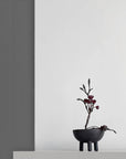 Coffee colour ceramic vase with branches and small flowers on a white surface against a gray wall.