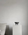 Dark grey colour bowl on a white pedestal against a plain gray wall with a gray sofa from 101 Copenhagen at Spacio retail stores in Mumbai & Kolkata.