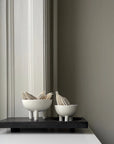 Two white ceramic bowls with plant stems on a black rectangular platform against a gray wall from 101 Copenhagen at Spacio retail stores in Mumbai & Kolkata.
