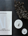 White ceramic jars with lids on a dark surface with 'Architectural Digest' magazine from 101 Copenhagen at Spacio retail stores in Mumbai & Kolkata.