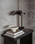 Dark metal desk lamp on a wooden surface with books and a bowl against a concrete wall.
