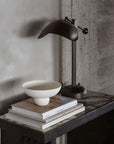 Dark metal desk lamp on a wooden surface with books and a bowl against a concrete wall.