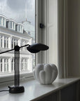 Modern black desk lamp on a windowsill with a white vase and building reflection.