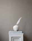 White sculpture of an apple with a feather on a gray pedestal against a gray wall from 101 Copenhagen at Spacio retail stores in Mumbai & Kolkata.