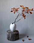 White vase with a branch and orange leaves on a gray background from 101 Copenhagen at Spacio retail stores in Mumbai & Kolkata.