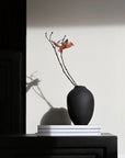 Black vase with a single branch on a white surface from 101 Copenhagen at Spacio retail stores in Mumbai & Kolkata.