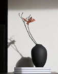Black vase with a single branch on a white surface from 101 Copenhagen at Spacio retail stores in Mumbai & Kolkata.