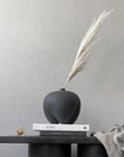 Black vase with dried plant on a table against a gray wall from 101 Copenhagen at Spacio retail stores in Mumbai & Kolkata.