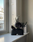 A black vase, known as the 101Cph Cobra Fat Medio Black 212004, gracefully sits on a window sill.