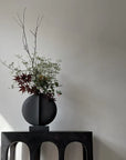 A quirky black vase, 101 Copenhagen Guggenheim Mini Coffee 111220, sits on a table in front of a white wall.