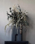 Decorative arrangement of dried plants in a black vase against a plain wall.