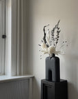 Black vase with white flowers on a small black pedestal (plinth) by a window