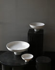 Three white ceramic bowls of varying sizes on a dark surface with a dark gray background from 101 Copenhagen at Spacio retail stores in Mumbai & Kolkata.