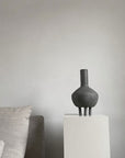 A 101Cph Duck Fat Dark Grey 111281 vase sits on a pedestal in front of a couch.