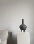 A 101Cph Duck Fat Dark Grey 111281 vase sits on a pedestal in front of a couch.