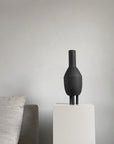 A 101Cph Duck Slim Coffee 111280 vase by 101 Copenhagen sits on a pedestal in front of a couch.