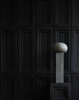 An architectural reference of a ceramic 101Cph Empire Big Dark Grey 223034 vase, resembling a concrete ball, elegantly sits on a pedestal in front of a black wall.