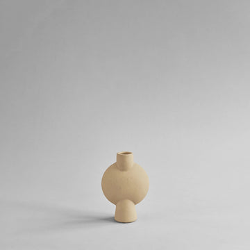 A beige vase with a sand finish sitting on a gray surface. This Scandinavian brand, 101 Copenhagen, offers the stylish 101Cph Sphere Bubl Mini Sand 203008 as an elegant home decor piece.