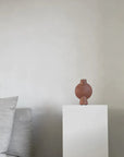 A brown 101Cph Sphere Bubl Mini Terracotta 203010 vase from the Scandinavian brand 101 Copenhagen sits on a white pedestal in a living room.
