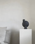 Black abstract sculpture on a white pedestal against a neutral wall with gray cushions from 101 Copenhagen at Spacio retail stores in Mumbai & Kolkata.
