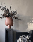 A 101 Copenhagen brand vase, the 101Cph Sphere Vase Square Big Terracotta 212008, with a Terracotta finish, placed on a table next to a chair.
