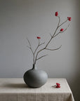 Gray vase with branches and red flowers on a beige tablecloth against a gray background from 101 Copenhagen at Spacio retail stores in Mumbai & Kolkata.