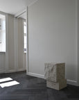 Tall limestone plinth in a minimalistic room with white walls and wooden floor.
