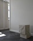 Tall limestone plinth in a minimalistic room with white walls and wooden floor.