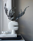 White vase with dried plants on a shelf styled with coffee table books against a gray wall from 101 Copenhagen at Spacio retail stores in Mumbai & Kolkata.
