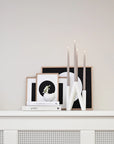 Decorative shelf with framed art, candles, and books against a plain wall.