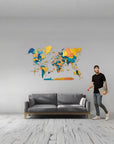 Man standing next to a gray sofa with a colorful large size world map on the wall behind him.