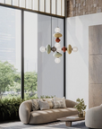 A modern multi-light pendant chandelier with colorful organic-shaped glass shades hangs in a contemporary living room with a beige sofa, brick walls, and large windows.