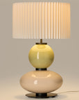 Lit up, modern table lamp with a yellow and white glass base and a pleated shade from Aromas. Available at Spacio retails stores in India.