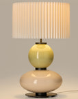 Lit up, modern table lamp with a yellow and white glass base and a pleated shade from Aromas. Available at Spacio retails stores in India.