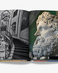 A grand, black-and-white staircase with ornate iron railings and intricate sculptures and a close-up of an ancient marble statue of a bearded man on a double spread in a luxurious Assouline Milan Chic coffee table book, showcasing Milan’s timeless elegance, now available at Spacio India’s online store and retail outlets.