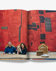 A stylish couple with their dog seated at a wooden dining table, set against a bold red and black textured artwork in an elegant interior on a double spread in a luxurious Assouline Milan Chic coffee table book, showcasing Milan’s timeless elegance, now available at Spacio India’s online store and retail outlets