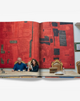 A stylish couple with their dog seated at a wooden dining table, set against a bold red and black textured artwork in an elegant interior on a double spread in a luxurious Assouline Milan Chic coffee table book, showcasing Milan’s timeless elegance, now available at Spacio India’s online store and retail outlets