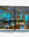 A double spread of a featuring mesmerizing display of suspended whale skeletons inside the Oceanographic Museum of Monaco, illuminated by soft blue lighting, highlighting the grandeur of marine life and history in a luxurious Assouline's Monte Carlo coffee table book, featuring a vibrant cover with images of Monaco's iconic landmarks, available at Spacio India.