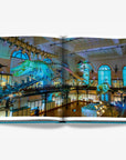 A double spread of a featuring mesmerizing display of suspended whale skeletons inside the Oceanographic Museum of Monaco, illuminated by soft blue lighting, highlighting the grandeur of marine life and history in a luxurious Assouline's Monte Carlo coffee table book, featuring a vibrant cover with images of Monaco's iconic landmarks, available at Spacio India.