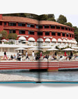 A seaside resort with red facade with waterfront terrace double spread in a luxurious Assouline's Monte Carlo coffee table book, featuring a vibrant cover with images of Monaco's iconic landmarks, available at Spacio India.