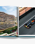 A double spread of the thrill of a left packed luxury apartment building with spectators watching the race, and on the right, a high-speed Red Bull Formula 1 car on the famous Monte Carlo street circuit in a luxurious Assouline's Monte Carlo coffee table book, featuring a vibrant cover with images of Monaco's iconic landmarks, available at Spacio India.