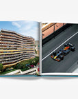 A double spread of the thrill of a left packed luxury apartment building with spectators watching the race, and on the right, a high-speed Red Bull Formula 1 car on the famous Monte Carlo street circuit in a luxurious Assouline's Monte Carlo coffee table book, featuring a vibrant cover with images of Monaco's iconic landmarks, available at Spacio India.