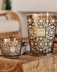 Two lit scented candles in decorative glass holders with a black geometric pattern over a warm cream background, displayed on a rustic wooden surface in a softly blurred indoor setting.